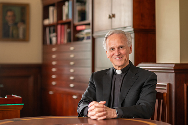 Priest with gray hair sitting at a wooden table, smiling at the camera.