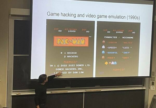 Luis Felipe Murillo points at a large classroom screen showing images of the title screens from the Pac-Man video game with certain words altered via hacks