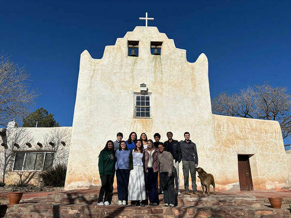 A group of ten students stands on the steps of a white adobe church with a bell tower under a clear blue sky. A light brown dog stands near the group.