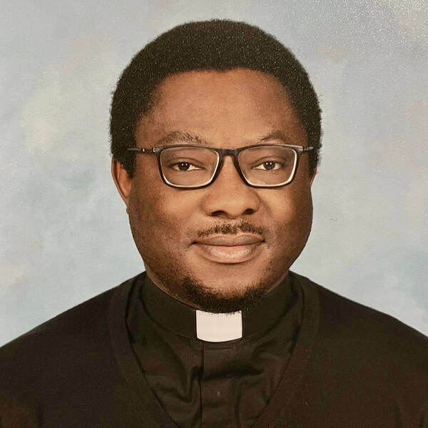 Headshot of a priest wearing glasses and a black clerical shirt with white collar against a light blue background.
