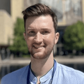 Man with short brown hair, light blue collared shirt, and lanyard smiles in front of Hesburgh Library.