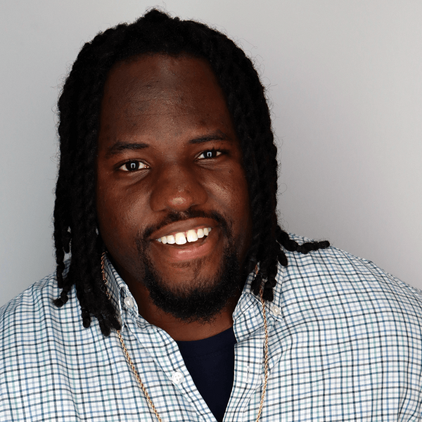 Headshot of a man with long, twisted black hair, wearing a blue-green plaid shirt over a dark blue undershirt, and a gold chain. He has a dark beard and is smiling broadly against a gray background.
