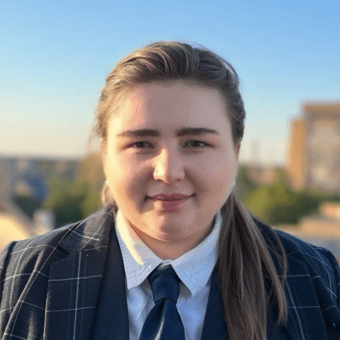 Headshot of a person with light brown hair pulled back, wearing a white collared shirt, navy blue tie, and dark green plaid blazer, against a blurred background of buildings and a clear blue sky.