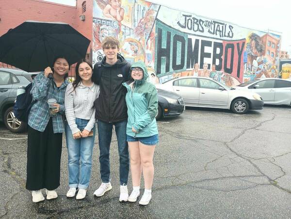 Four smiling young people, three females and one male, stand under a black umbrella. The group includes a woman in plaid, a man in a black jacket, and a woman in a teal rain jacket. Behind them is a colorful mural on a brick building that reads "JOBS not JAILS HOMEBOY." Wet street.