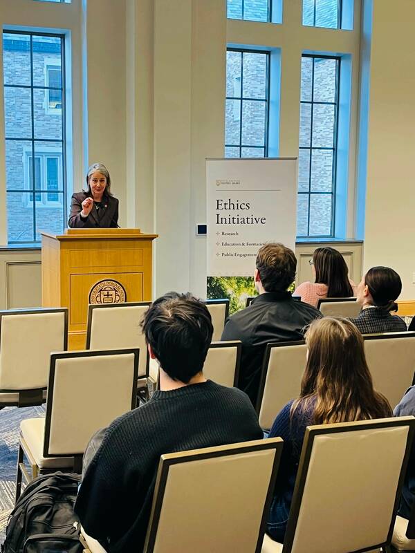 A woman in a brown blazer gestures while speaking at a podium to an audience at a Notre Dame Ethics Initiative event.