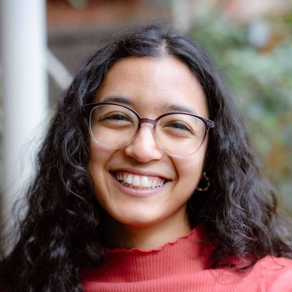 A young woman with dark curly hair and glasses smiles broadly, wearing a coral ruffled shirt.