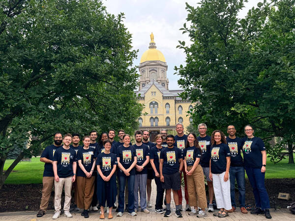 Smiling group in "Summer of Love Camp" shirts pose before the Golden Dome of Notre Dame's Main Building.