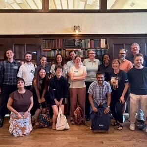 A diverse group of 20+ smiling people, some kneeling, pose in a wood-paneled room lined with full bookshelves.