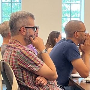 Three men, one with a gray beard and plaid shirt, others in a blue tee and plaid, sit attentively at a table.