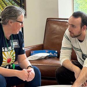 A woman with glasses and a man with a beard sit conversing. The woman wears a nametag; a brown couch is behind them.