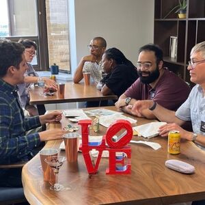 Six diverse people converse at wooden tables in an office. A red "LOVE" sculpture sits prominently on the foreground table.