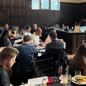 A man speaks at a podium to a seated audience in a formal wood-paneled dining room with drinks and food on tables.