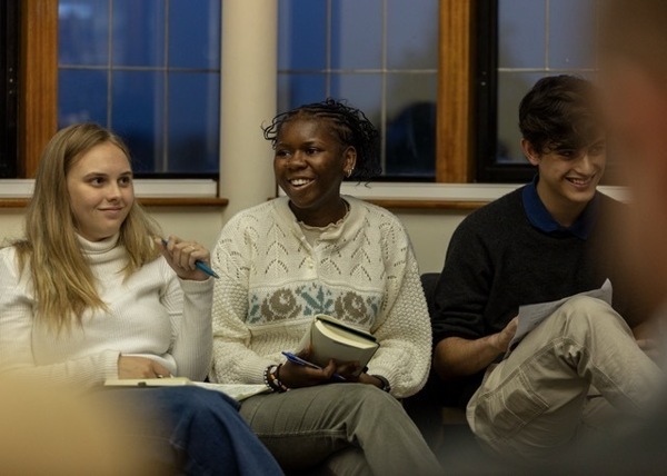 Three Notre Dame students smile and are attentive in a room. A woman in a white patterned sweater holds a notebook.