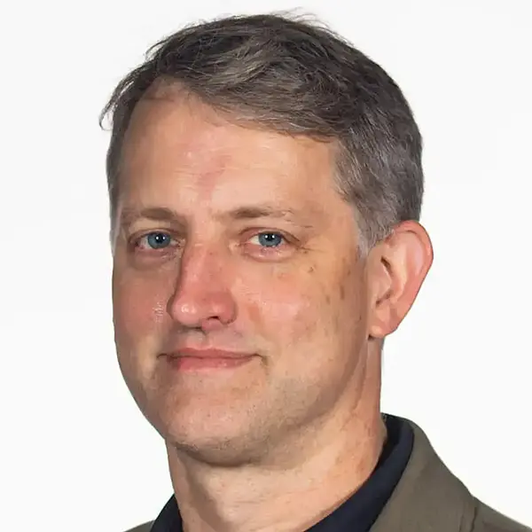 Headshot of a smiling man wearing a light brown jacket over a dark blue shirt.