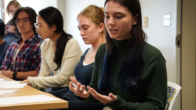 A student with dark hair and a blue streak, wearing a green turtleneck, speaks with gesturing hands at a classroom table.