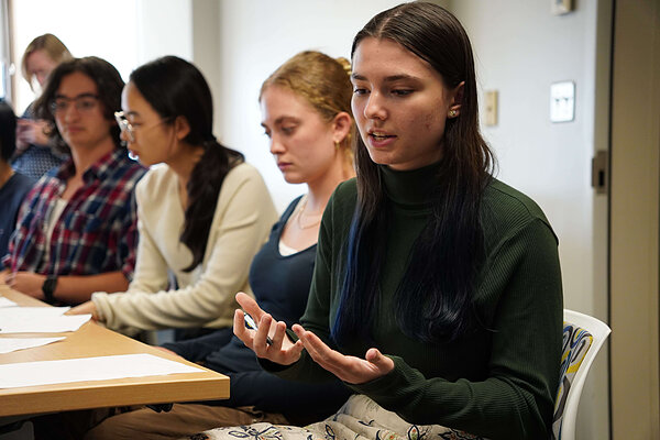 A student with dark hair and a blue streak, wearing a green turtleneck, speaks with gesturing hands at a classroom table.